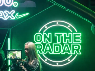 Green neon “On The Radar” sign glowing on a dark wall beside a Nike Air Max logo, with ogpriness by a camera rig in the foreground under moody studio lighting