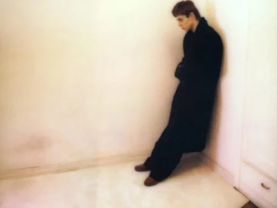 A softly blurred portrait of The Kid LAROI leaning into a corner, dressed in a long dark coat against a pale, minimal room, conveying isolation and quiet emotional restraint aligned with the tone of “I Condemn.”