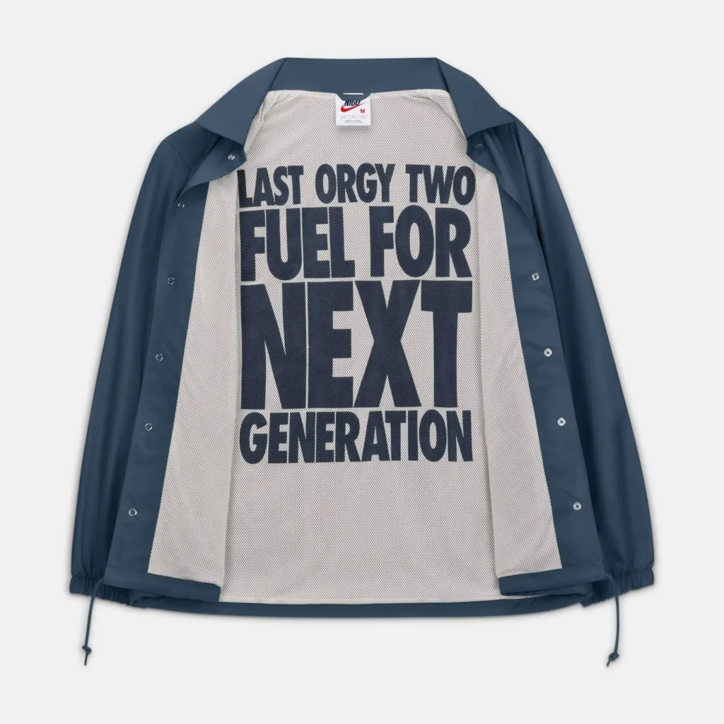 Navy snap-button coach jacket displayed open to reveal a contrasting white mesh lining printed with bold oversized typography reading “LAST ORGY TWO / FUEL FOR NEXT / GENERATION.” Elasticated cuffs and hem with drawstrings frame the relaxed silhouette, while the minimal exterior contrasts with the graphic-heavy interior design