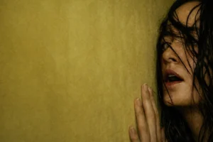 A distressed Renate Reinsve presses her hand against a yellow, fluorescent-lit wall, her damp hair partially obscuring her face as she looks upward with a tense, disoriented expression in a liminal interior setting