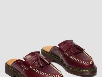 A pair of burgundy leather mule loafers shown from a front three-quarter angle, featuring a backless slip-on design. The shoes highlight a polished finish with tassel detailing across the vamp and bold white contrast stitching outlining the moc-toe. The tan suede footbed is partially visible, while the chunky black sole with signature yellow welt stitching adds a sturdy, distinctive foundation