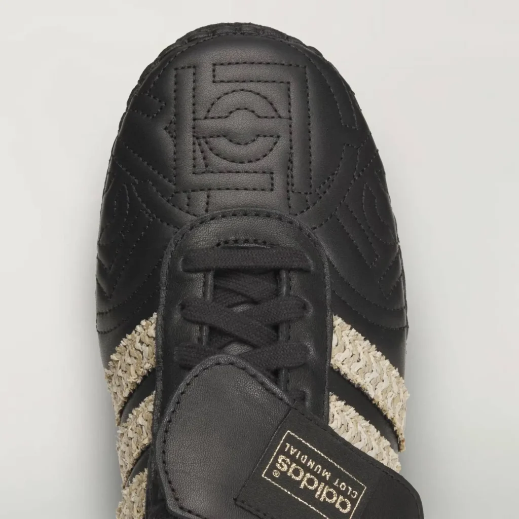 top-down close-up of black adidas x CLOT Mundial shoe showing quilted leather upper, tonal stitched monogram detail, lace system, and raffia-woven Three Stripes with branded tongue label