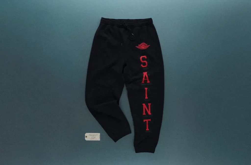 Black SAINT Mxxxxxx x Jordan Brand sweatpants displayed flat against a muted grey backdrop, featuring bold red vertical “SAINT” lettering along the leg, an embroidered red wing logo on the upper thigh, and an elastic waistband with drawstring