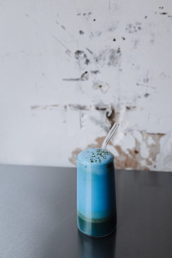 Tall cylindrical glass filled with a layered blue beverage—deep teal at the base fading into a lighter sky-blue foam on top—sprinkled with green powder and served with a clear glass straw, set on a dark table against a textured, weathered wall with peeling paint