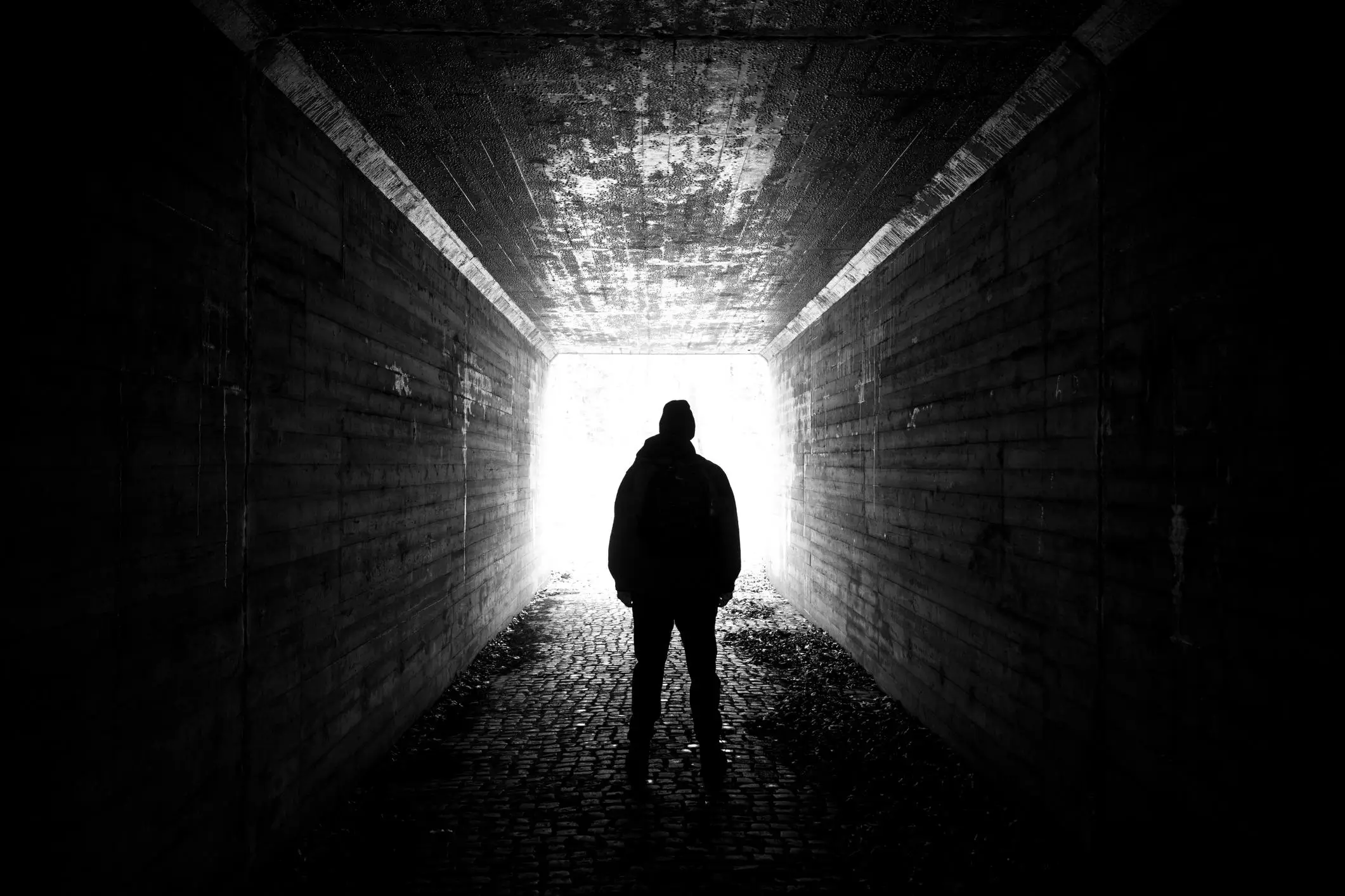 A lone figure stands silhouetted inside a dark concrete tunnel, facing a blinding white exit. The person appears to be wearing a hooded jacket and a backpack, their outline sharply contrasted against the light. The tunnel’s rough, weathered walls and cobblestone ground add a gritty, urban texture, while the strong backlighting creates a stark, cinematic mood—suggesting isolation, transition, or a quiet moment before stepping into the unknown