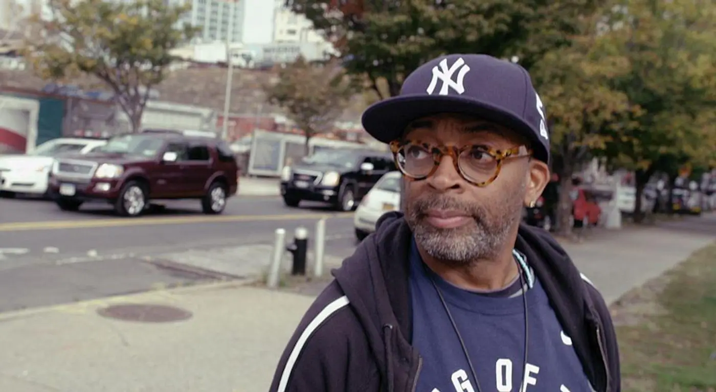 Spike Lee walking along a city street wearing a New York Yankees cap, round tortoiseshell glasses, and a dark hoodie, glancing to the side with cars and urban background softly blurred behind him