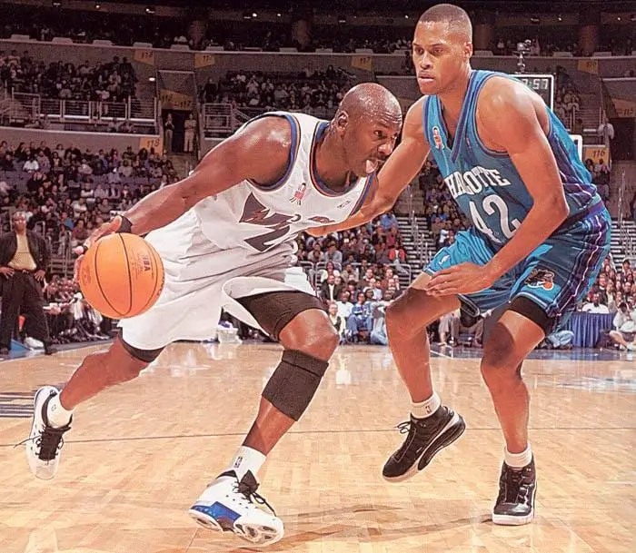 Michael Jordan, wearing a white Washington Wizards uniform, drives low to the basket while dribbling with his left hand, closely guarded by a Charlotte Hornets defender in a teal jersey on a hardwood NBA court, with a packed arena crowd blurred in the background