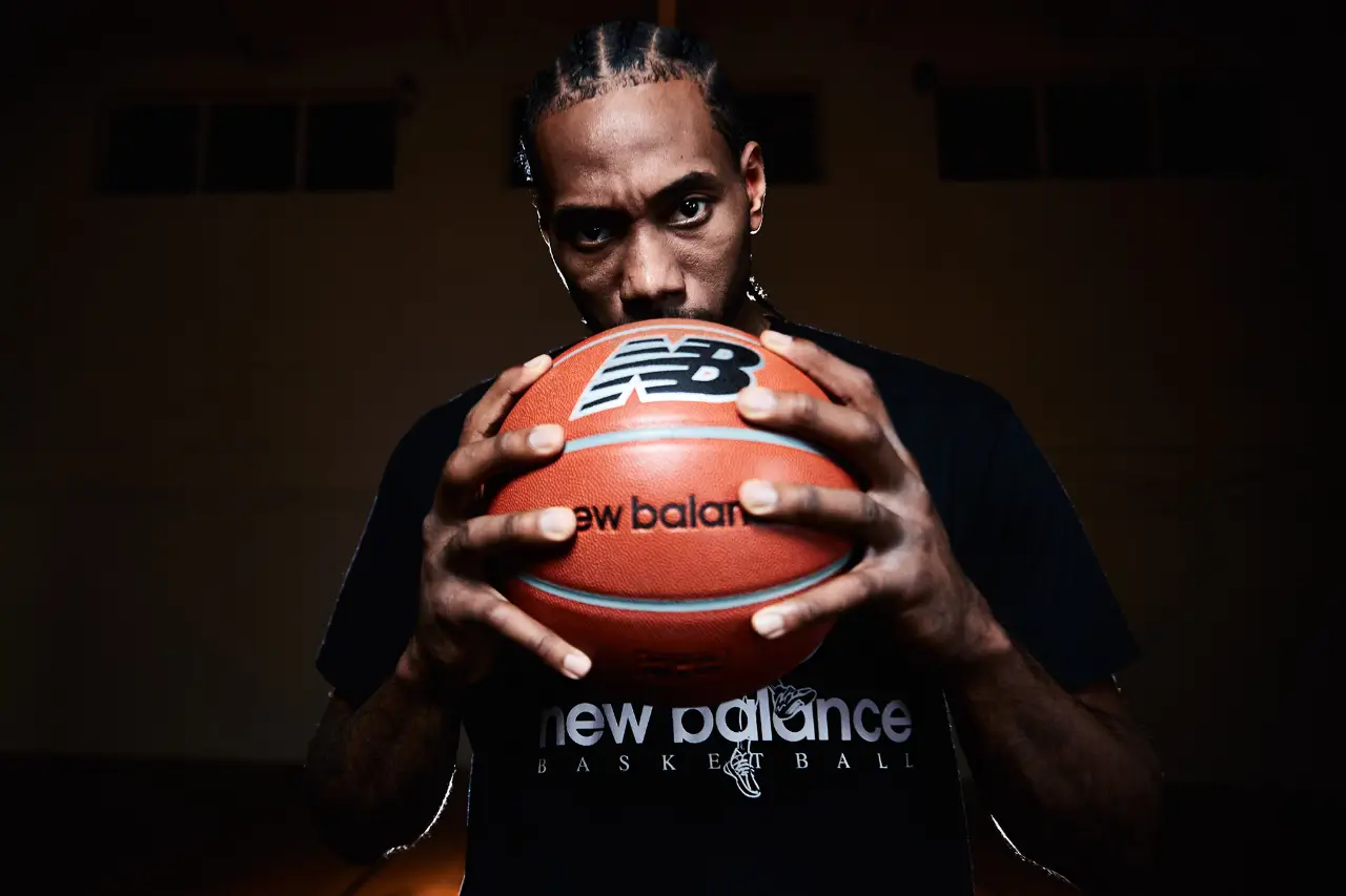 Kawhi Leonard holding a New Balance basketball in a dark gym setting, wearing a New Balance Basketball shirt and staring intensely at the camera