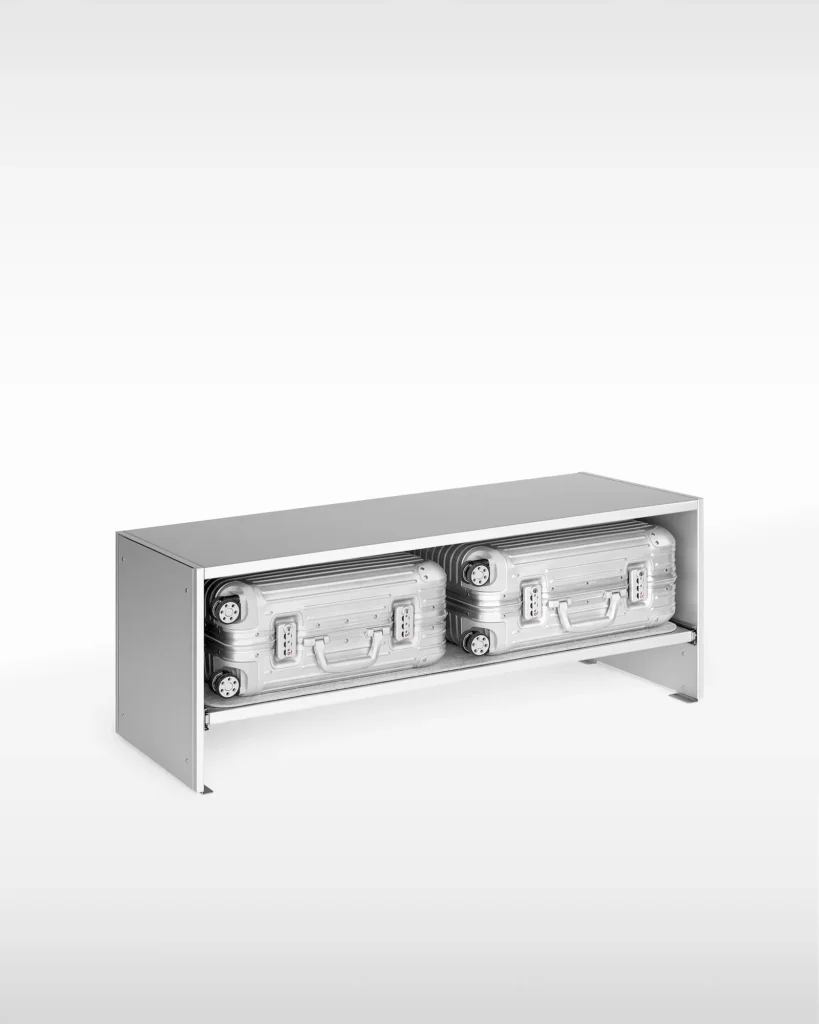 Minimalist light gray metal storage bench with an open front, housing two compact hard-shell suitcases side by side on a pull-out shelf against a clean white background