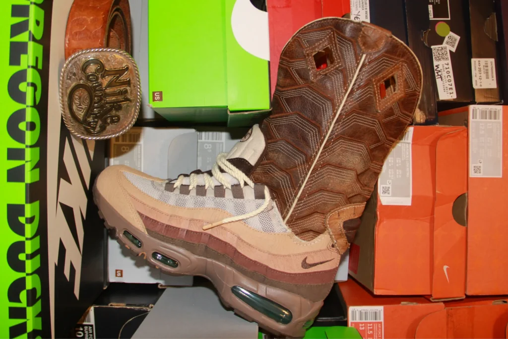 Custom Nike Air Max 95 transformed into a cowboy boot with a stitched leather shaft, displayed against stacked shoe boxes and Western accessories