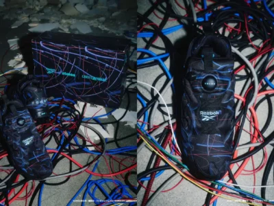 A pair of futuristic black sneakers with neon blue and red abstract line graphics are displayed among a chaotic spread of tangled cables on a concrete floor. One image shows the shoes beside a matching shoebox branded with Reebok, atmos, and Ghost in the Shell, while the other highlights a top-down view of the shoe, featuring a central circular detail near the tongue, a pull tab, and visible Reebok branding. The cyberpunk aesthetic is reinforced by the surrounding wires and dim, industrial setting