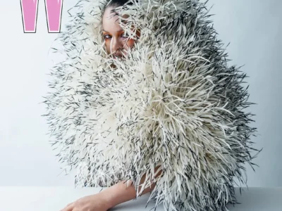 A magazine-style cover image featuring a Rihanna wrapped in an oversized, textured feather-like garment in white with subtle black accents, creating a voluminous, cocooned silhouette. Only part of her face is visible through the layers, with bold eye makeup peeking through. Her hand extends forward on a clean, minimal surface, showcasing long, pointed black nails. The background is soft and neutral, while large typography includes a pink “W” at the top and the words “The Pop Issue,” with “Rihanna” displayed prominently in bright orange at the bottom