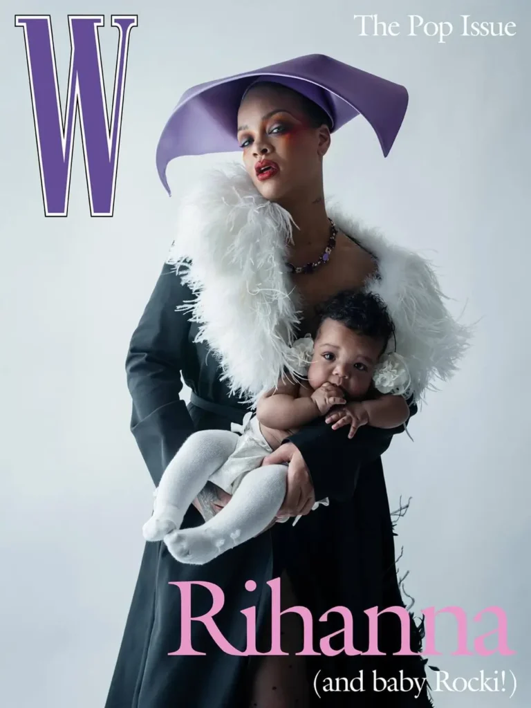A magazine cover-style portrait of a woman holding a baby against a clean, minimal backdrop. She wears a sculptural lavender hat, a black coat with a dramatic white feathered collar, and bold makeup with warm-toned eyeshadow and red lips. The baby, dressed in soft white with tights, rests in her arms, adding a tender contrast to the high-fashion styling. Large typography includes a purple “W” at the top, “The Pop Issue,” and “Rihanna (and baby Rocky!)” in pink lettering, reinforcing the editorial, cover-ready composition