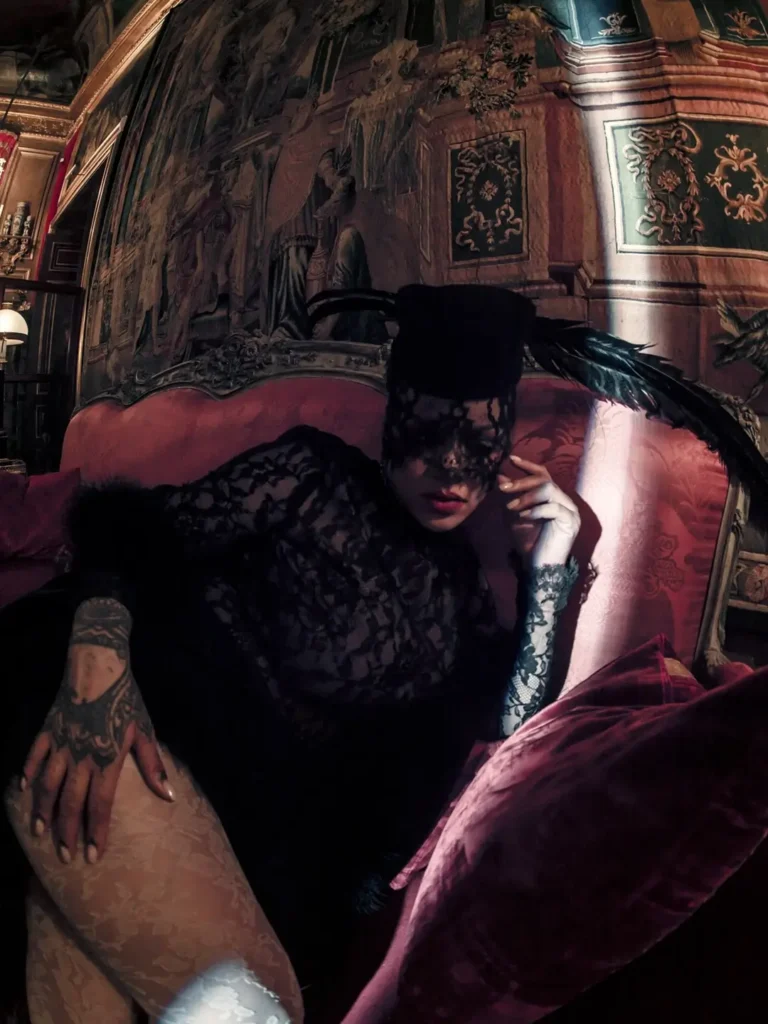 A moody, cinematic fashion portrait set in an ornate, baroque-style interior, where a woman reclines on a plush red velvet sofa. She wears a sheer black lace outfit with long sleeves and a matching lace veil that partially obscures her face, paired with a structured black headpiece adorned with a feather. Her pose is languid and composed, one hand resting on her leg, revealing intricate tattoos. Rich textures—velvet, lace, and painted wall panels—combine under low, dramatic lighting to create an atmosphere of dark elegance and intimate theatricality