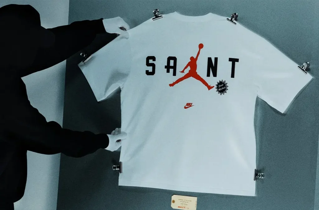 White SAINT Mxxxxxx x Jordan Brand T-shirt clipped to a display wall, featuring bold “SAINT” lettering across the back with a red Jumpman silhouette integrated into the design, a small Nike logo below, and a connective graphic badge, held in place by gloved hands against a muted grey backdrop