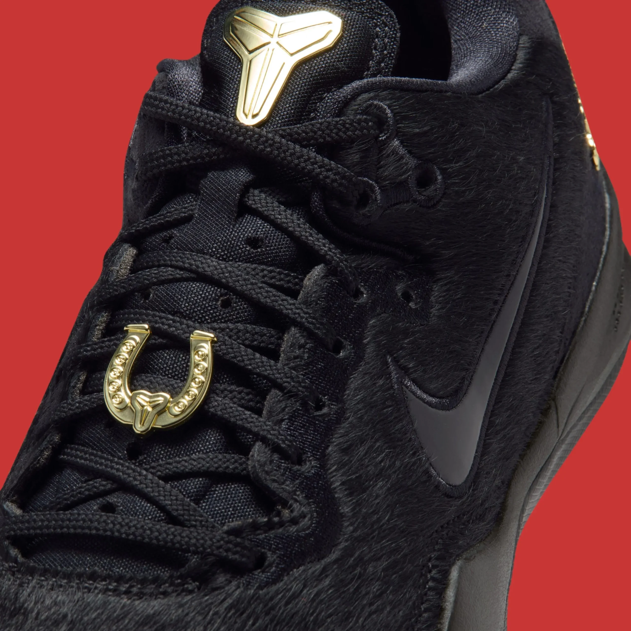 Close-up of a black Nike basketball shoe featuring a textured, fur-like upper with black laces and a padded tongue. A metallic gold Kobe logo sits prominently at the top of the tongue, while a gold horseshoe-shaped lace charm with embossed detailing anchors the laces below. A dark Swoosh is partially visible along the side, set against a bold red background that heightens the contrast and haute feel