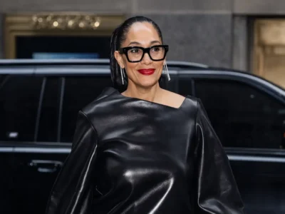 Tracee Ellis with slicked-back hair wearing bold black rectangular eyeglasses and a structured black leather top, paired with bright red lipstick, standing outdoors in front of a dark vehicle at night
