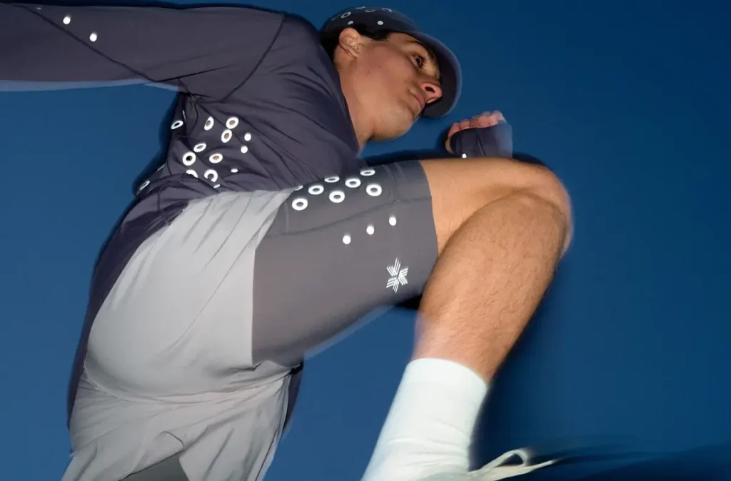 A man is captured mid-movement against a vivid blue sky, his body angled diagonally as if in the stride of a run or a dynamic stretch. One knee is lifted high while his opposite arm is bent forward, creating a sense of motion and athletic tension. He wears a coordinated performance outfit: a fitted, long-sleeve top and two-tone running shorts in shades of gray