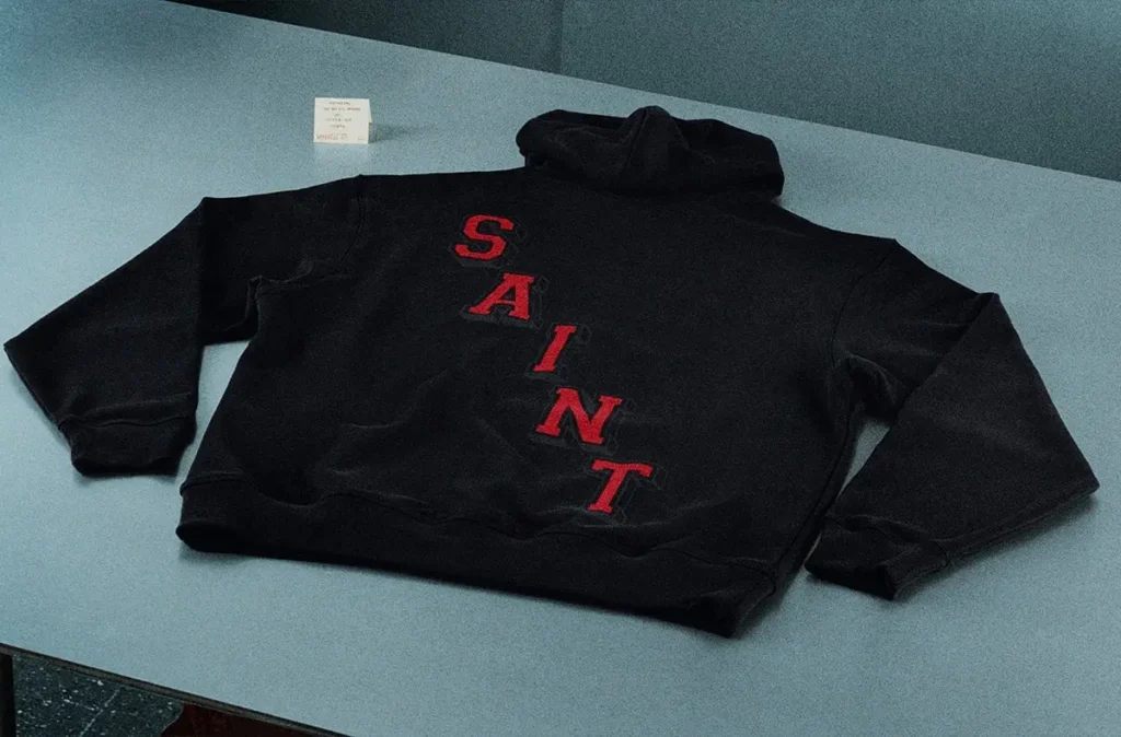 Black SAINT Mxxxxxx x Jordan Brand hoodie laid flat on a display surface, featuring bold red “SAINT” lettering running diagonally across the back, with a relaxed silhouette and minimal detailing against a muted grey backdrop