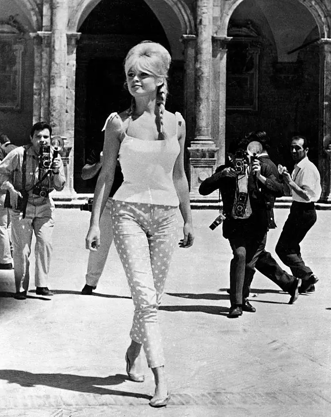 Black-and-white photograph of Brigitte Bardot walking confidently through a courtyard in a fitted top and patterned trousers, surrounded by photographers capturing her movement, reflecting her status as a 1960s media sensation