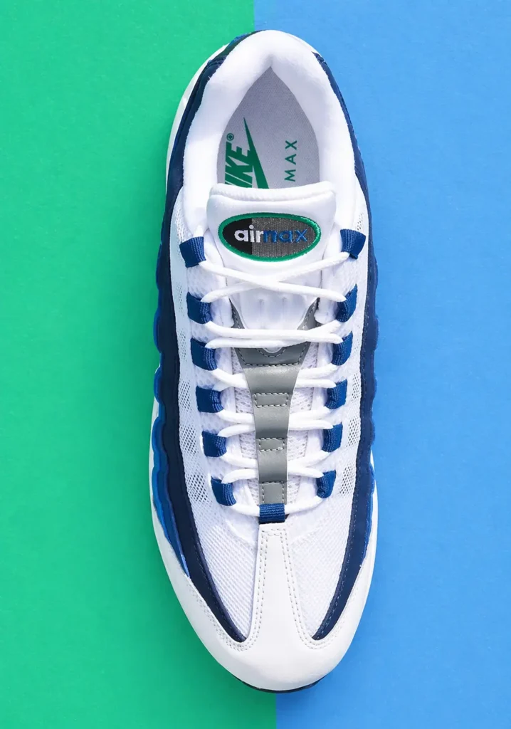 Top-down view of a Nike Air Max 95 sneaker in white with navy and blue accents, centered on a two-tone ground green and blue back