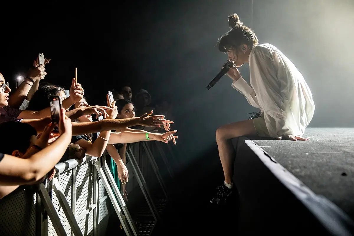 Noga Erez seated at the edge of the stage mid-performance, leaning toward the crowd while holding a microphone, as fans reach out with phones raised, capturing the intimate concert moment under dramatic spotlighting