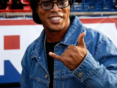 Ronaldinho black beret and glasses wearing a blue denim jacket, making a relaxed hand gesture in a stadium setting with a blurred crowd in the background