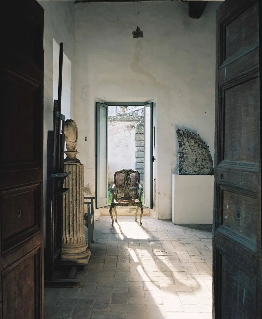 A sunlit, aged interior corridor frames an ornate wooden chair placed directly in an open doorway, with light casting long shadows across stone flooring; classical sculpture fragments and architectural elements line the walls, reinforcing a quiet, museum-like atmosphere