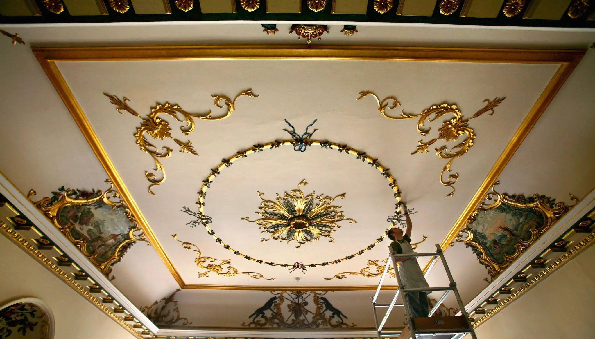 Artisan standing on a scaffold restoring an ornate ceiling, featuring intricate gilded scrollwork, painted panels, and a central decorative medallion within a historic interior