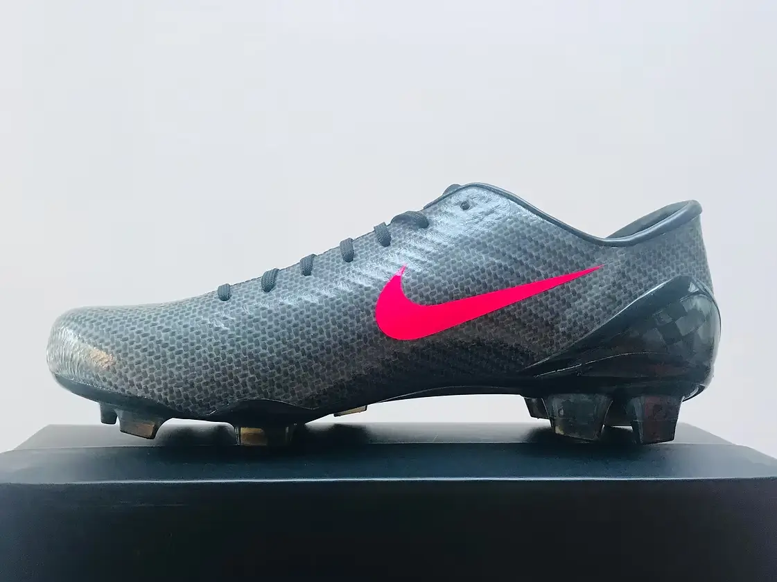 Sleek low-profile football boot in a dark textured synthetic finish, featuring a bright pink Swoosh, molded studs, and a streamlined silhouette displayed on a minimal surface