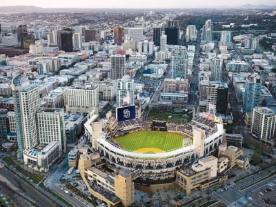Aerial view of downtown San Diego centered on Petco Park, with the baseball field visible inside the stadium, surrounded by modern high-rise buildings, dense city blocks, and distant hills under a hazy sky
