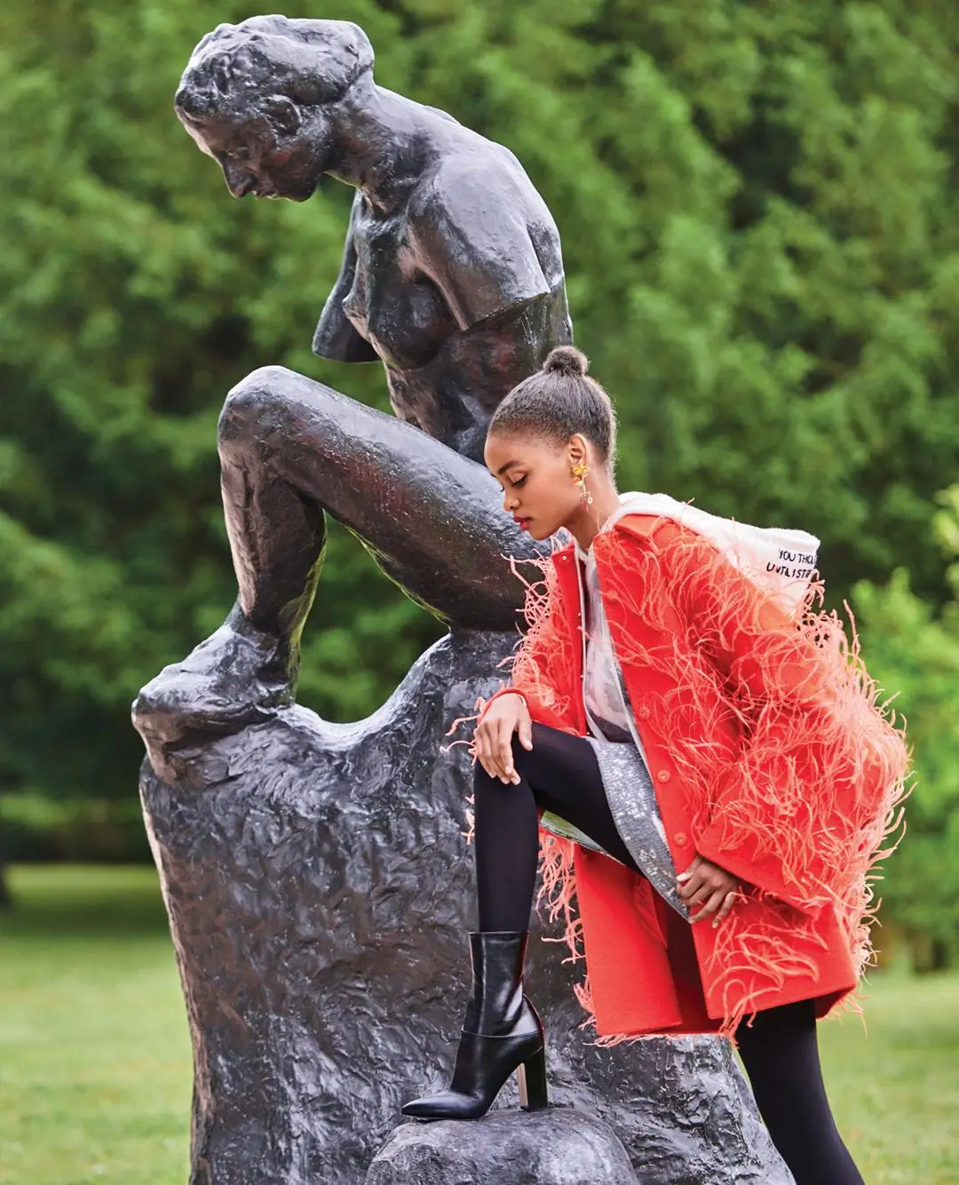 A fashion image of a model posing beside a dark bronze sculpture of a seated figure in a lush outdoor setting, wearing a vibrant red coat with feathered detailing, black leggings, and heeled boots, with one foot placed on the sculpture’s base in a poised, editorial stance