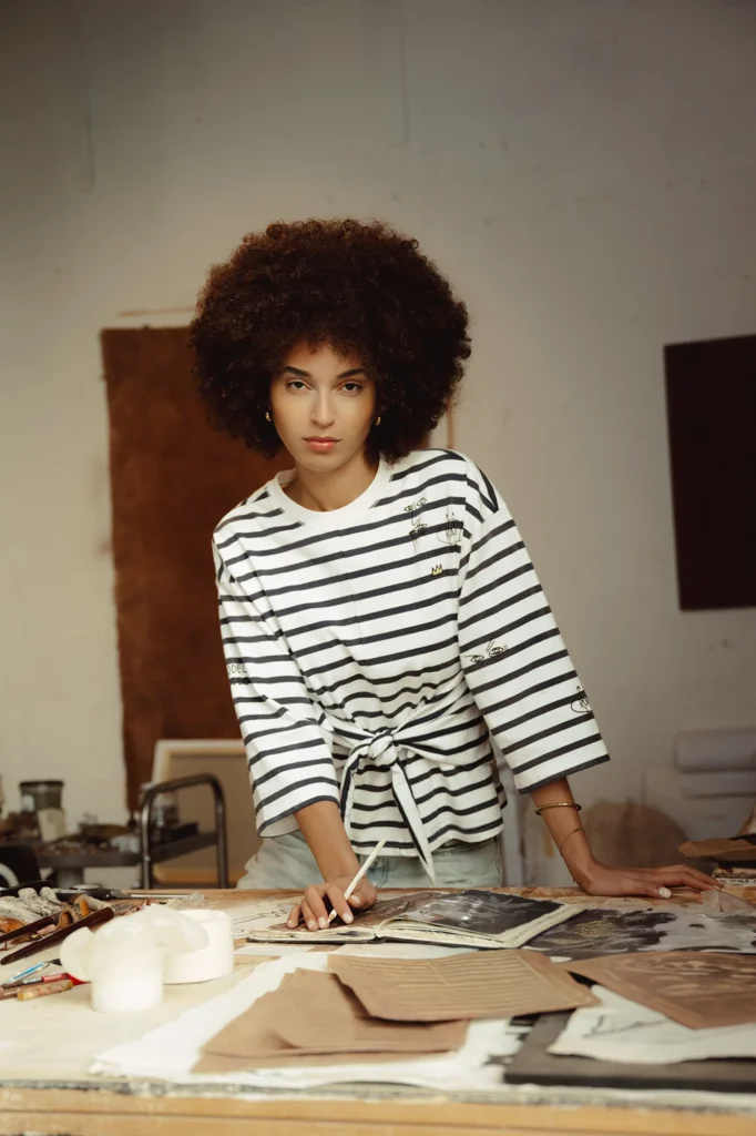 A woman with a voluminous natural afro stands leaning over a wooden worktable in an artist’s studio, looking directly at the camera with a composed, confident expression. She wears a loose, black-and-white striped top with three-quarter sleeves and a tied waist detail, paired with light-colored bottoms. Her hands rest on an open sketchbook or magazine spread, surrounded by scattered papers, textured materials, and art tools. The workspace feels tactile and in-progress, with neutral-toned canvases or boards leaning against the wall in the background, soft natural lighting, and a quiet, creative atmosphere that suggests design, illustration, or mixed-media work in motion