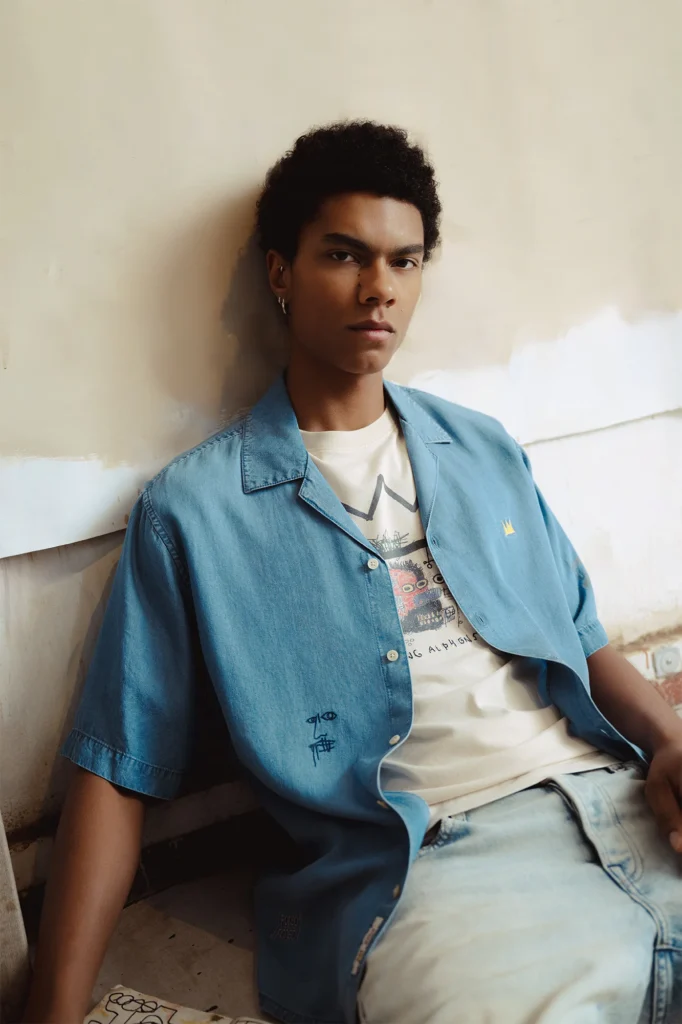 A young man with short, curly hair leans casually against a textured studio wall, gazing directly at the camera with a calm, self-assured expression. He wears a light blue short-sleeve button-up shirt layered over a graphic T-shirt featuring Basquiat-inspired artwork, paired with light-wash jeans. Subtle embroidered details appear on the shirt, adding an artistic accent. The setting feels raw and minimal, with partially painted walls and soft natural lighting, creating an understated, contemporary atmosphere that blends fashion with creative studio energy