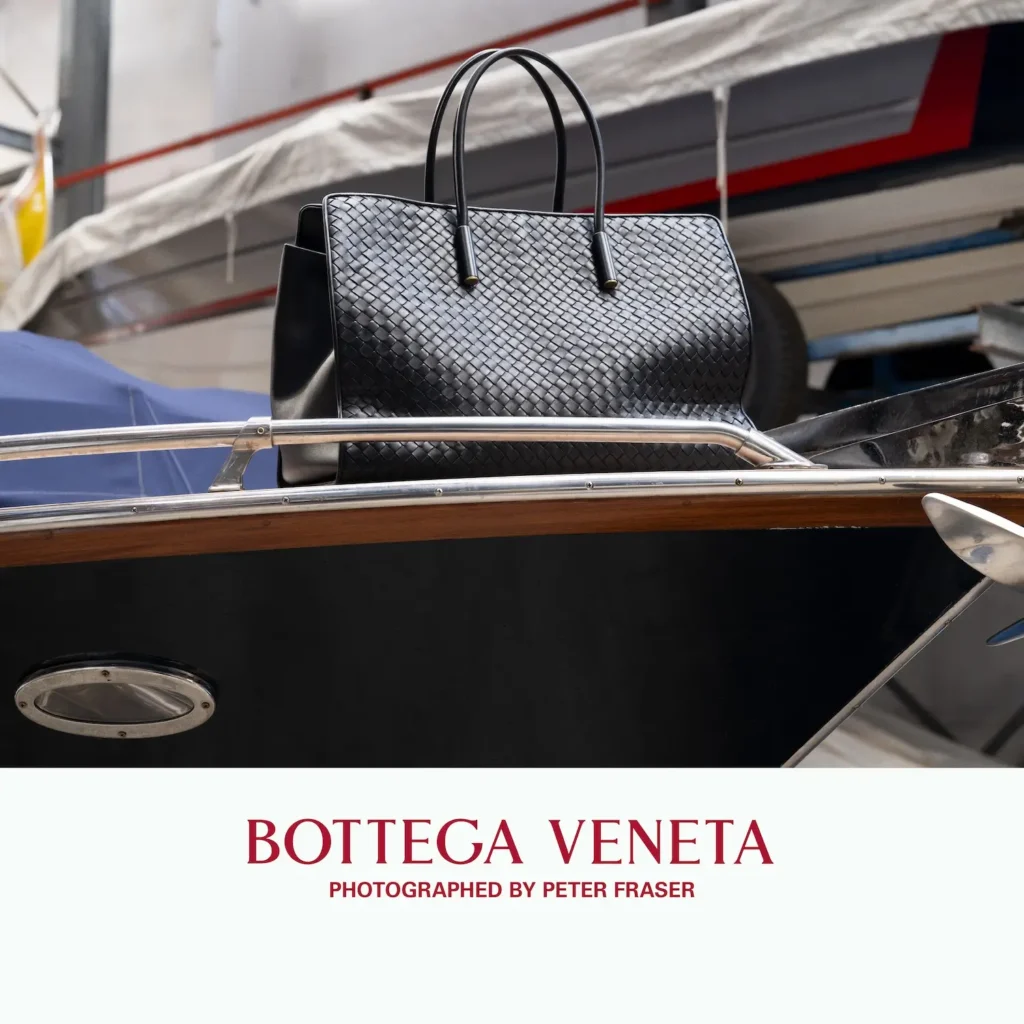 Black woven leather tote bag by Bottega Veneta with structured handles, displayed atop a polished wooden speedboat in an industrial marina setting; campaign text below reads “Bottega Veneta, Photographed by Peter Fraser.”