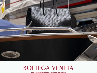 Black woven leather tote bag by Bottega Veneta with structured handles, displayed atop a polished wooden speedboat in an industrial marina setting; campaign text below reads “Bottega Veneta, Photographed by Peter Fraser.”