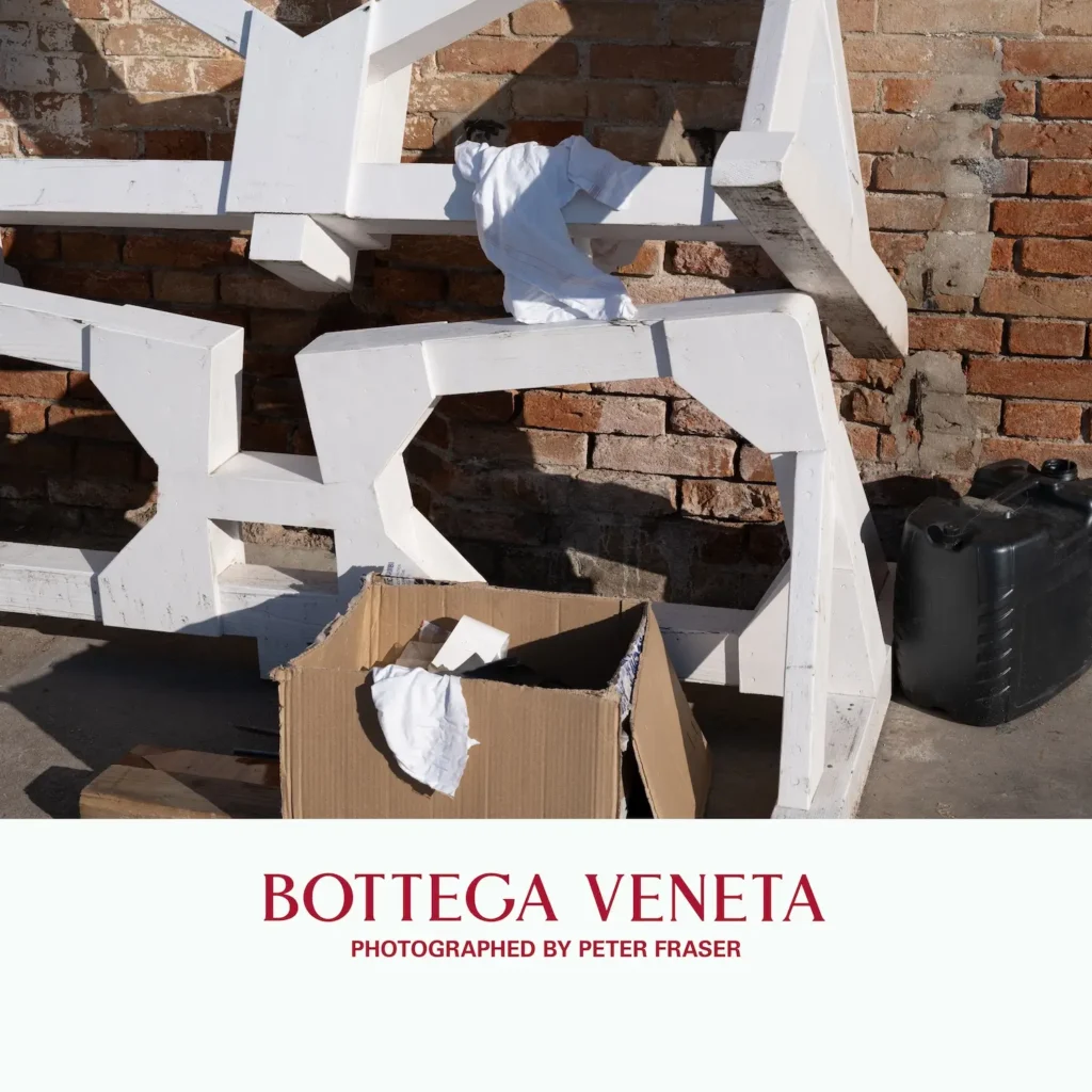 Abstract white wooden structures arranged against a brick wall with cloths draped over them, a cardboard box and containers nearby suggesting a workshop or outdoor workspace; campaign text below reads “Bottega Veneta, Photographed by Peter Fraser.”
