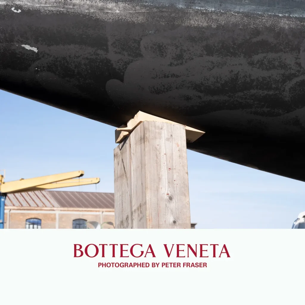 Large boat hull supported by a wooden post in an outdoor industrial setting, with a building and crane visible in the background; campaign text below reads “Bottega Veneta, Photographed by Peter Fraser.”