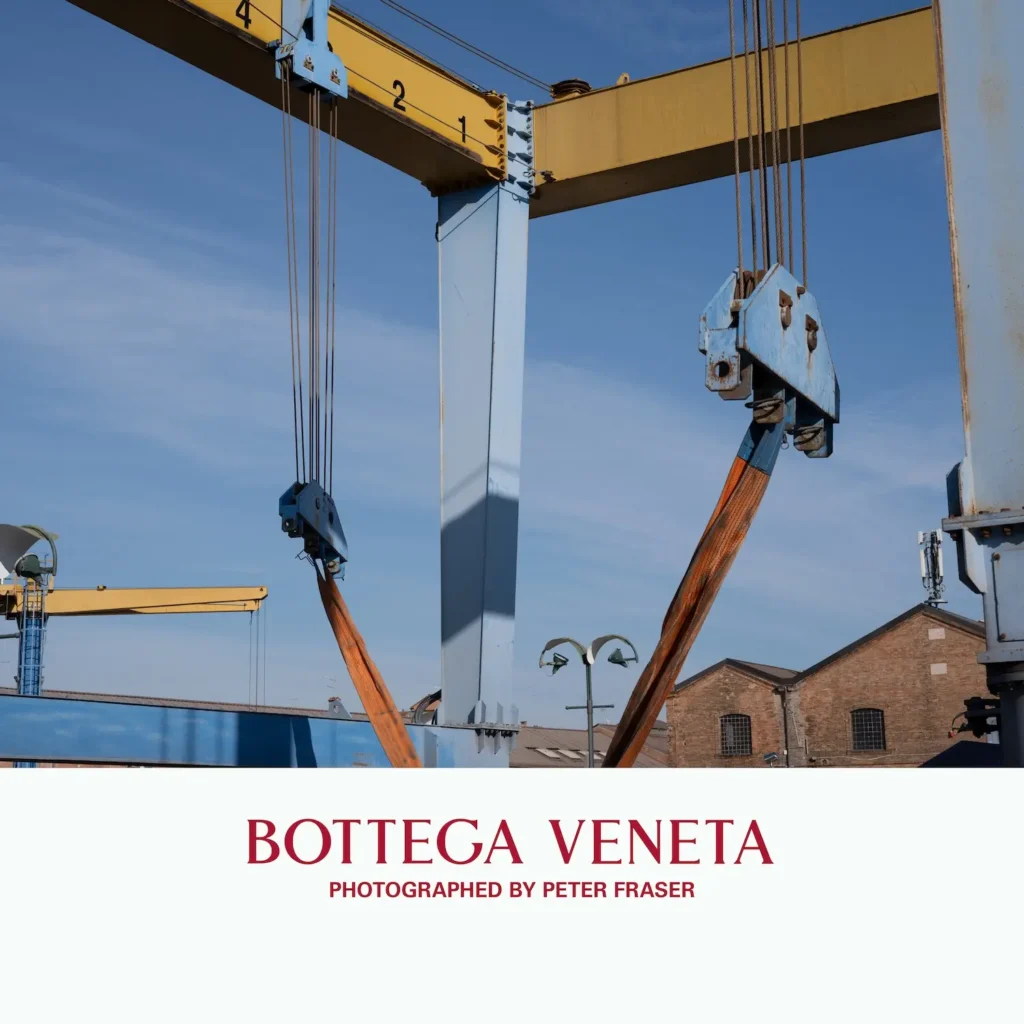 Industrial crane structure with suspended lifting straps and metal hooks set against a blue sky, with brick buildings in the background; campaign text below reads “Bottega Veneta, Photographed by Peter Fraser.”
