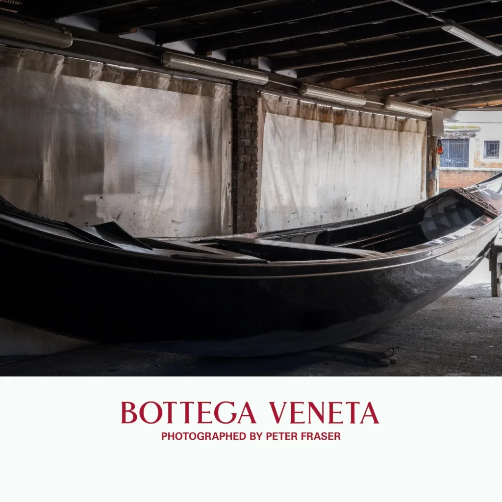 Glossy black boat hull resting inside a covered workshop space with exposed beams and translucent curtains filtering light; campaign text below reads “Bottega Veneta, Photographed by Peter Fraser.”
