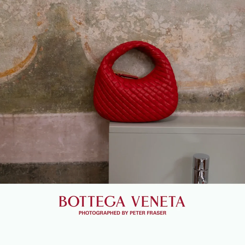 Red woven leather handbag with a rounded silhouette placed on a minimalist pedestal against a weathered textured wall; campaign text below reads “Bottega Veneta, Photographed by Peter Fraser.”