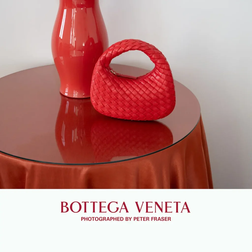 Red woven leather handbag with a rounded handle displayed on a glossy round table beside a matching red vase, creating a tonal still-life composition; campaign text below reads “Bottega Veneta, Photographed by Peter Fraser.”