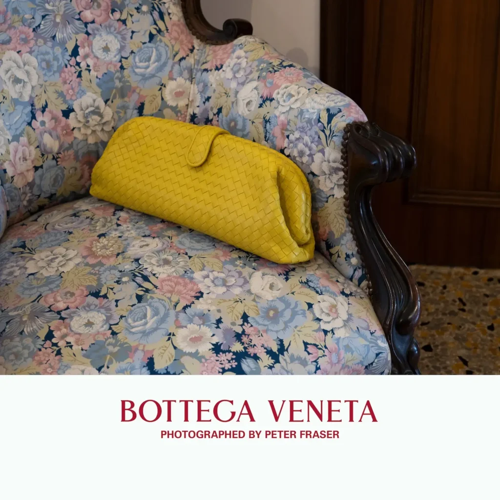 Yellow woven leather clutch resting on a vintage floral-upholstered armchair with carved wooden arms in a classic interior setting; campaign text below reads “Bottega Veneta, Photographed by Peter Fraser.”