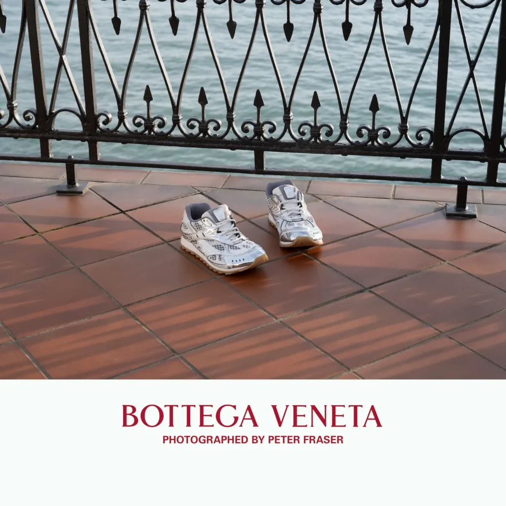 Pair of white technical shoe placed on terracotta tiles beside an ornate black iron railing overlooking water, lit by soft natural light; campaign text below reads “Bottega Veneta, Photographed by Peter Fraser.”