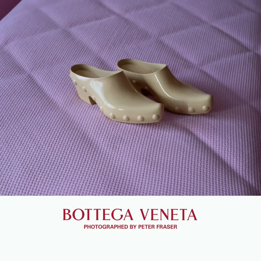 Pair of beige rubber clogs with rounded stud details, set on a textured pink fabric surface under soft directional light; campaign text below reads “Bottega Veneta, Photographed by Peter Fraser.”