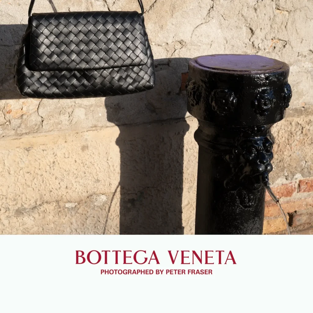 Black woven leather shoulder bag hangs against a textured stone wall beside a glossy black outdoor fountain, casting sharp shadows in sunlight; campaign text below reads “Bottega Veneta, Photographed by Peter Fraser.”