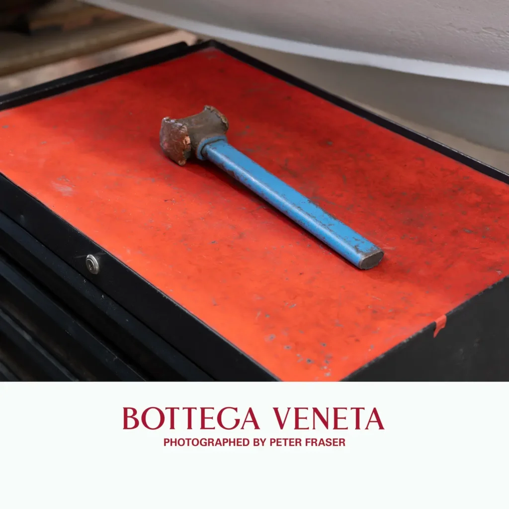 Worn metal hammer with a blue handle resting on a scratched red tool chest surface, suggesting a workshop environment; campaign text below reads “Bottega Veneta, Photographed by Peter Fraser.”