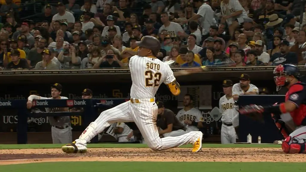 A dynamic in-game baseball moment showing a San Diego Padres batter Juan Soto wearing a white pinstriped uniform with “Soto 22” on the back, mid-swing as he drops to one knee after making contact, while the catcher in red gear reaches out behind him and a packed crowd watches from the stands
