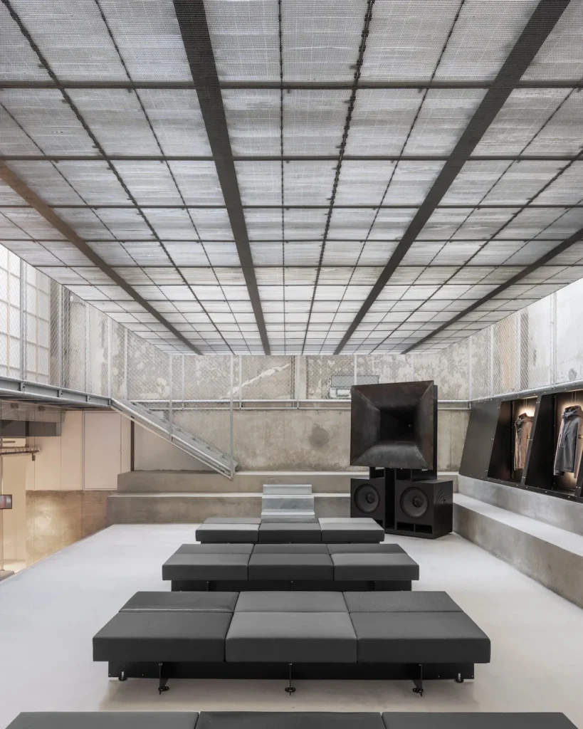 A minimalist, industrial-style interior space with tiered black modular seating arranged in rows facing a large sculptural speaker installation. The room features raw concrete walls, a translucent grid ceiling that diffuses soft light, and metal mesh paneling along the upper perimeter. A staircase ascends along the left side, while recessed wall displays on the right showcase hanging garments, blending elements of a gallery, showroom, and immersive listening environmen