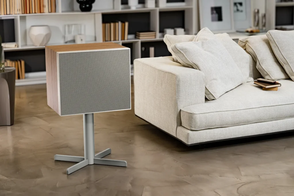 Geneva Lab DeCon/XL speaker in a clean white finish on a minimalist stand, centered in a moody modern interior with deep red ambient lighting, a lounge sofa, and sculptural furnishings creating a cinematic, design-forward atmosphere